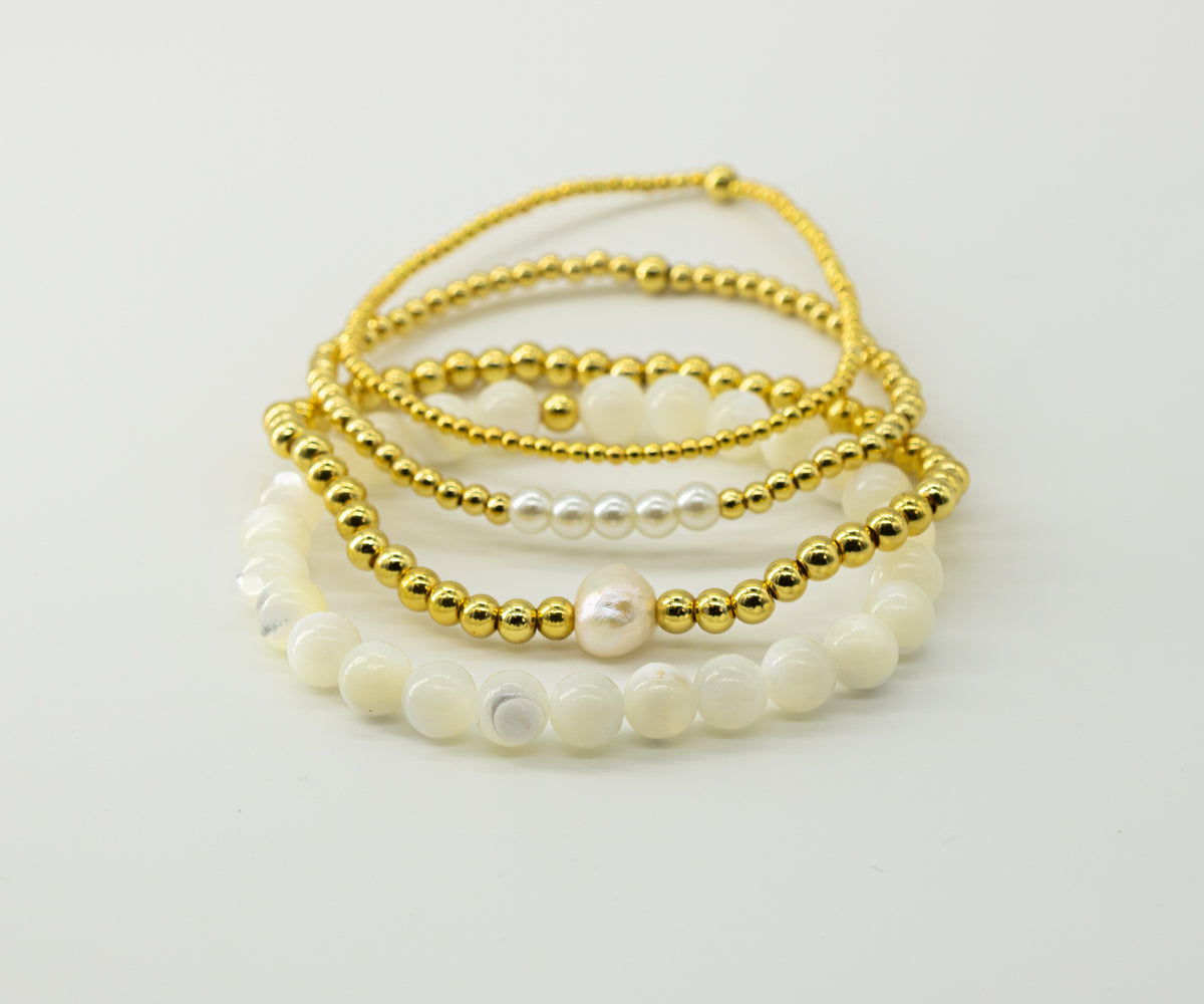 A stack of four handmade beaded bracelets on a clean white background made by evrgold. The top  bracelet is made of 18k gold filled 2mm beads. the next one is 18k gold filled 3mm beads with 5 glass faux white pearl beads as an accent. the next one is 4mm 18k gold filled beads with one accent genuine pearl bead. the bottom bracelet is made of all white mother of pearl beads.