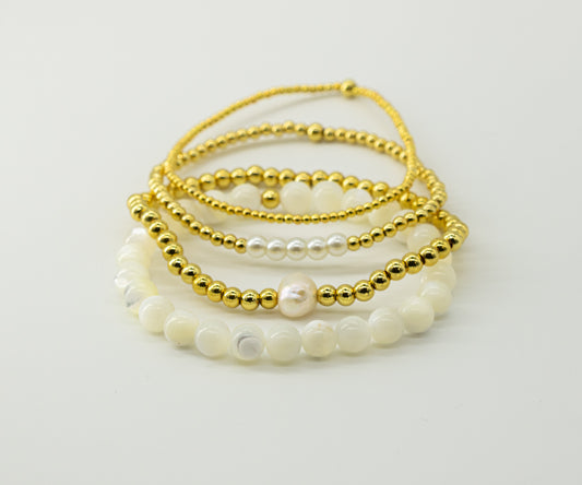 A stack of four handmade beaded bracelets on a clean white background made by evrgold. The top  bracelet is made of 18k gold filled 2mm beads. the next one is 18k gold filled 3mm beads with 5 glass faux white pearl beads as an accent. the next one is 4mm 18k gold filled beads with one accent genuine pearl bead. the bottom bracelet is made of all white mother of pearl beads.