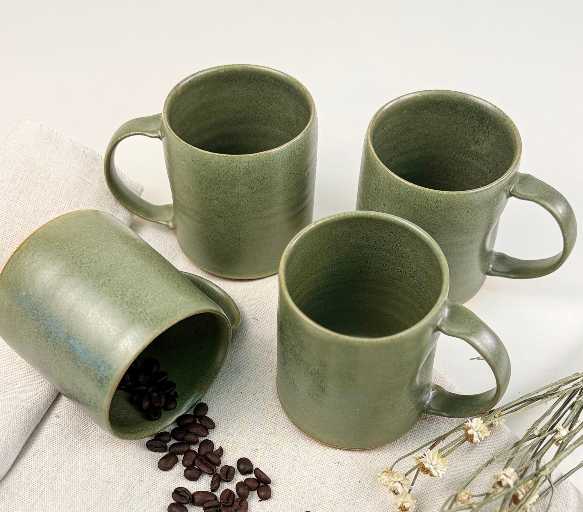 Four handmade sage green mugs styled flatly with coffee beans and dried flowers.